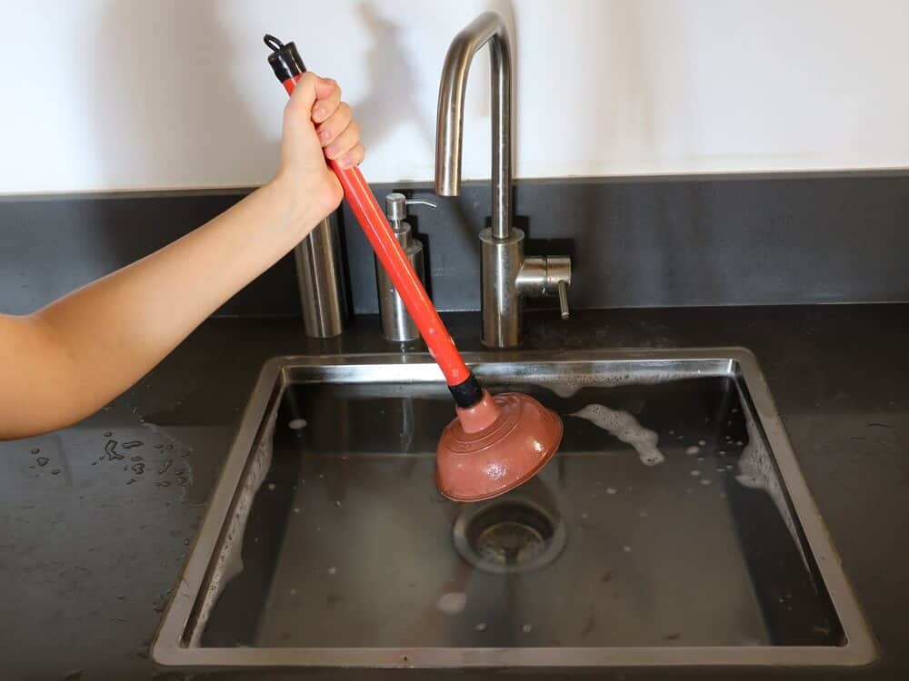 hand holding plunger on top of oveflowing kitchen sink with blocked drain